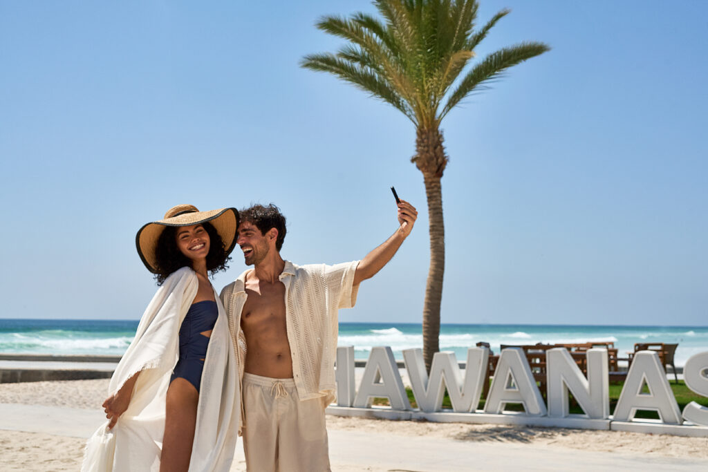 Happy couple taking a selfie on a sunny beach with Hawana Salalah sign and palm tree in the background at Juweira Boutique Hotel Hawana Salalah Oman