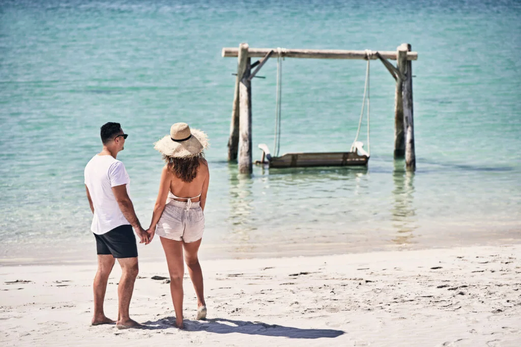 Couple holding hands on a sandy beach looking at a wooden swing over shallow turquoise waters at Fanar Hotel & Residences Hawana Salalah Oman